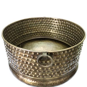 May include: A round, antique brass-colored metal planter with a hammered texture. The planter has a wide, open top, a decorative ring handle, and a raised base. The surface is covered in small, evenly spaced dimples.