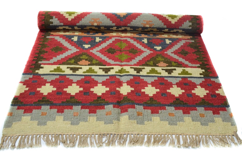 May include: A red, green and beige patterned rug with fringe. The rug has a geometric design with a central diamond shape.