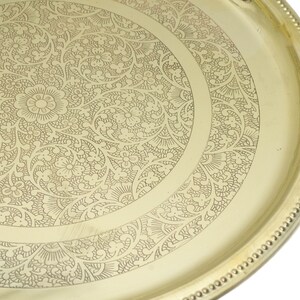May include: A gold-colored, round tray with an intricate floral and leaf pattern etched into the surface. The tray's edge features a beaded border. The design is centered with a large flower.