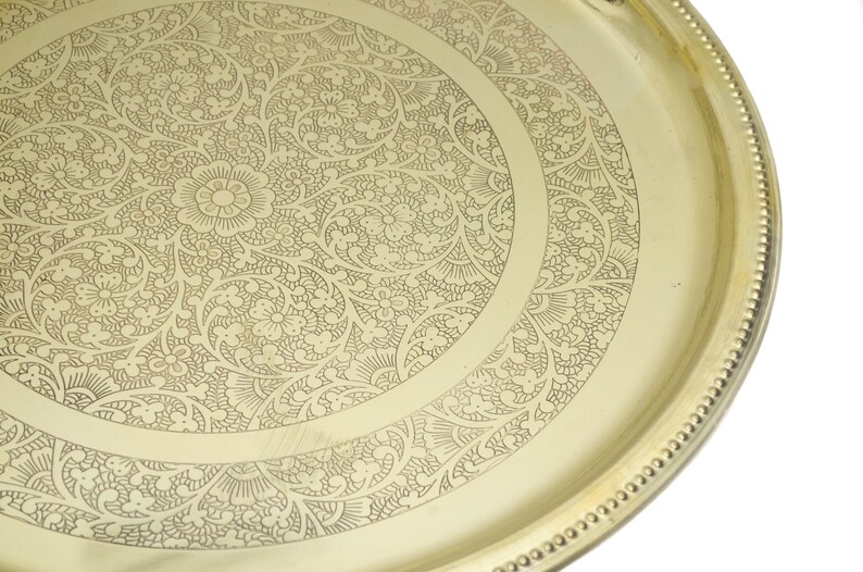 May include: A gold-colored, round tray with an intricate floral and leaf pattern etched into the surface. The tray's edge features a beaded border. The design is centered with a large flower.