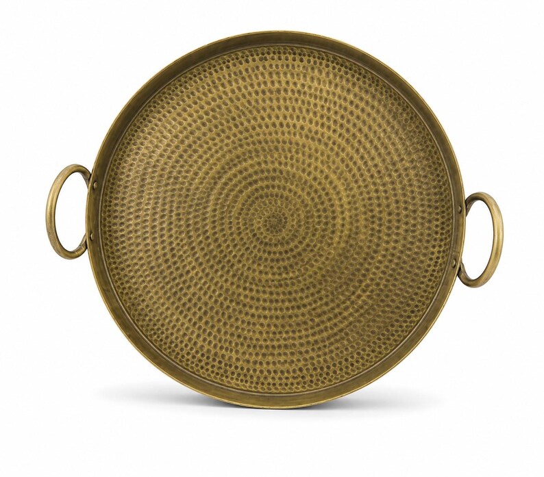 May include: A round brass tray with a hammered texture featuring a spiral pattern across the surface. The tray has a raised rim and two circular handles attached to opposite sides, providing a metallic finish with a warm golden-brown color.