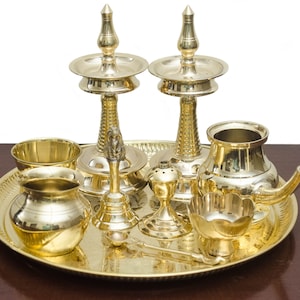 May include: A collection of brass religious items, including two ornate lamps, a small pot, a bowl, a water vessel with a spout, and other decorative pieces. The items are arranged on a round, golden tray.
