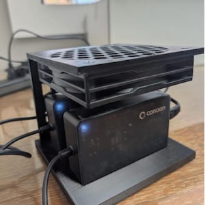 以下が含まれることがあります： 3Dプリントされた黒い冷却スタンドに、2台の長方形のCanaan製電源ユニットが収められています。スタンドの上部には六角形のグリッドカバーが付いた冷却ファンが取り付けられています。各電源ユニットには、小さな青いインジケーターライトが点灯しています。