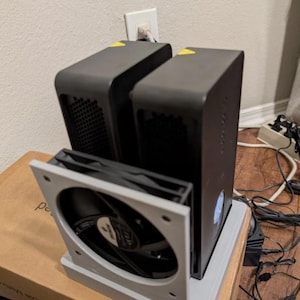 May include: Two rectangular dark gray electronic devices positioned vertically side-by-side, supported by a light gray plastic stand featuring a circular cooling fan mounted on the front. Small yellow warning stickers are visible on the top edges.