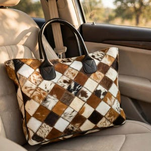 May include: A tote bag featuring a patchwork design of cowhide squares in shades of brown, white, and black. The bag has two black leather handles attached with triangular stitched tabs at the top edge, creating a structured rectangular shape.