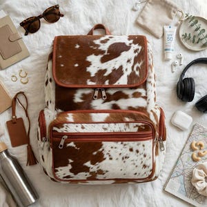 May include: A cowhide print backpack with brown and white patches, featuring a top flap, a front zippered pocket, and brown leather-like trim. The backpack is surrounded by various personal items including sunglasses, a notebook, headphones, and a water bottle.