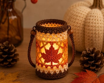 May include: A cylindrical glass candle holder encased in a crocheted sleeve featuring a dark brown rim and base with a cream and orange maple leaf pattern. The holder includes two dark brown crocheted handles attached with small wooden beads.