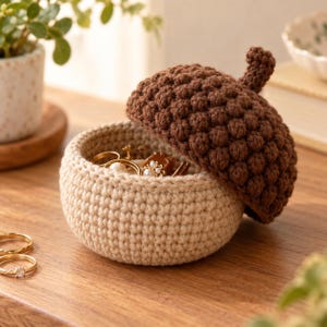 Puede incluir: Un joyero de ganchillo con forma de bellota, con una tapa texturizada de color marrón oscuro y una base de color beige claro. La tapa está entreabierta, dejando ver varios anillos dorados con pequeñas perlas blancas y piedras.