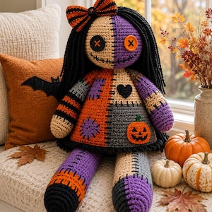 Può includere: Una bambola all'uncinetto fatta a mano con un viso bicolore beige e viola, occhi a bottone neri e una bocca cucita. La bambola indossa un vestito patchwork arancione, viola e grigio con un'applicazione a zucca, un cuore e un fiocco a righe.