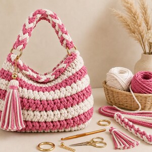 Op de afbeelding: Een gehaakte handtas met horizontale strepen van crème en magentakleurig garen. De tas heeft een gevlochten handvat bevestigd met goudkleurige ringen en een bijpassende kwast. Ernaast liggen garenbollen, een haaknaald, scharen en extra koord.