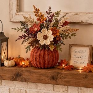 Pode incluir: Um vaso de abóbora em crochê preenchido com um buquê de flores e folhagens de outono em crochê nos tons creme, laranja, vermelho e verde. A abóbora é canelada e texturizada, servindo como base para o arranjo floral com diversas folhas.