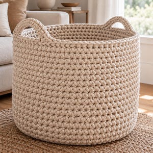 Puede incluir: Una cesta grande y redonda de ganchillo en color beige, con un patrón de punto grueso y texturizado y dos asas integradas en el borde superior. La cesta está hecha de un material similar a un cordón continuo, creando una estructura densa y uniforme.