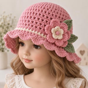 Op de afbeelding: Een gehaakte roze bucket hat met een geschulpte rand en een decoratieve gelaagde bloemapplicatie aan de zijkant. De bloem bestaat uit roze bloemblaadjes met een crèmekleurig hartje, vergezeld door twee kleine groene gehaakte blaadjes.