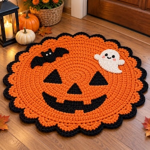 Puede incluir: Una alfombra redonda de ganchillo naranja con una cara de calabaza negra, un murciélago negro y un fantasma blanco. La alfombra tiene un borde negro festoneado. El diseño es plano, circular y está elaborado con puntadas de hilo visibles.