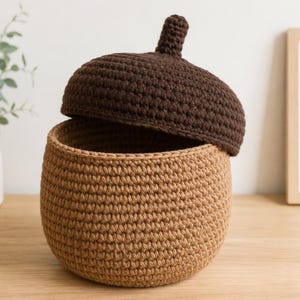 May include: A round, crocheted storage basket with a tan base and a dark brown, acorn-shaped lid. The basket features a textured, tight stitch pattern throughout, with the lid topped by a small, rounded knob resembling an acorn stem.