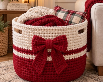 May include: A round, two-toned crochet basket featuring a cream-colored upper section and a deep red lower section. A large, matching red crochet bow is attached to the front. The basket has integrated side handles and contains soft textiles.