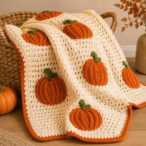 May include: A cream-colored crochet blanket featuring a repeating pattern of raised, textured orange pumpkins with green stems. The blanket has a solid orange border and is draped over a woven basket, showcasing a detailed handmade knit design.