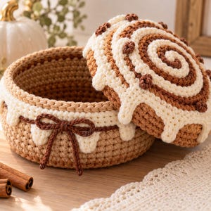 May include: A round, crocheted storage basket with a lid designed to resemble a cinnamon roll. The basket features tan, cream, and dark brown yarn with a decorative dark brown bow on the side and cream-colored icing-like trim.