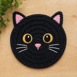 May include: A circular black crochet coaster shaped like a cat face. The design features two pointed ears with pink inner details, large yellow eyes with black pupils, a small pink nose, and three white embroidered whiskers on each side.