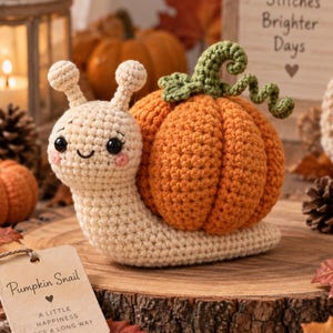 May include: A cream-coloured crochet snail with black eyes and pink cheeks, featuring a large orange pumpkin shell with a green curly stem. A tan tag attached to the snail reads: Pumpkin Snail, A little happiness goes a long way.