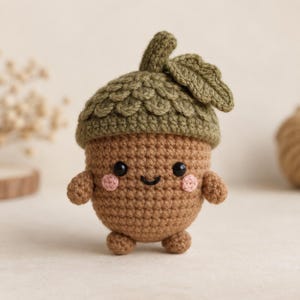Puede incluir: Un juguete de peluche de bellota tejido a ganchillo con cuerpo marrón y una tapa verde oliva texturizada con una pequeña hoja. La figura tiene dos ojos de cuentas negras, una sonrisa negra bordada y dos pequeñas mejillas rosadas, con brazos y pies.