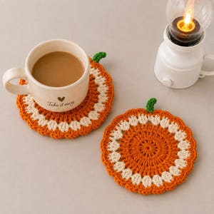 May include: Two round crochet coasters shaped like pumpkins, featuring orange and cream yarn with a small green stem. One coaster supports a beige ceramic mug with the text "Take it easy" and a heart icon printed on the side.