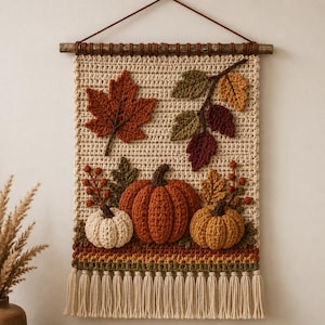 May include: A rectangular crochet wall hanging featuring a cream-colored background with three-dimensional autumn motifs including a large orange pumpkin, a smaller white pumpkin, a golden pumpkin, red maple leaves, and brown tassels at the bottom.