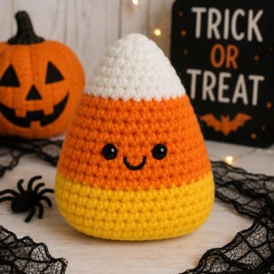 May include: A crochet candy corn plush toy featuring a white top, orange middle, and yellow bottom section. The plush has two small black plastic eyes and a stitched black smile. The texture is made of visible yarn loops in a triangular shape.