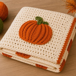 May include: A folded cream-colored crochet blanket featuring a textured orange pumpkin appliqu&eacute; with a green stem. The blanket has a grid-like stitch pattern and an orange border, showcasing a repeating pumpkin motif along the folded edges.