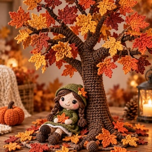 May include: A crocheted doll with brown braided hair wearing a green hooded dress sits under a large crocheted tree with orange, red, and yellow maple leaves. A small crocheted pumpkin and scattered maple leaves surround the base of the tree.