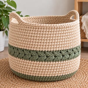 May include: A round, hand-crocheted storage basket made of thick beige yarn with two integrated handles. The basket features a wide horizontal band of dark green yarn with a textured, braided stitch pattern and a solid dark green base.