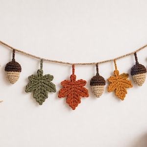 Puede incluir: Una guirnalda decorativa con hojas de arce y bellotas de ganchillo en tonos naranja, amarillo, marrón y verde, suspendidas de un cordel trenzado. Los elementos están dispuestos en un patrón alterno a lo largo de la cuerda.