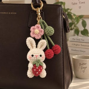 Peut inclure: Un porte-clés en crochet représentant un lapin crème tenant une fraise rouge, agrémenté d'une fleur rose, d'une sphère verte et de deux cerises rouges, fixé à un mousqueton en métal doré suspendu à un sac à main marron foncé.