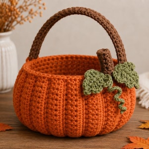 May include: A round, orange crochet basket shaped like a pumpkin, featuring a brown handle, a brown stem, and two green leaves with a curly vine attached to the side. The basket has a ribbed texture and is constructed from yarn stitches.