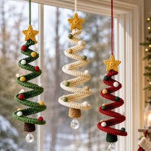 Op de afbeelding: Drie spiraalvormige kerstboomhangers van haakwerk die verticaal hangen. Elke boom heeft een gele ster aan de bovenkant, kleine kleurrijke pomponversieringen, een bruine basis en een heldere gefacetteerde kristallen kraal onderaan.