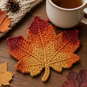 May include: A crochet maple leaf coaster featuring a gradient colour pattern that transitions from deep red at the top to bright orange and golden yellow at the base, with visible stitched veins radiating from the centre stem to the leaf edges.