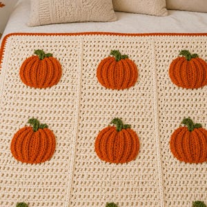 May include: A cream-colored crochet blanket featuring a repeating pattern of textured orange pumpkins with green stems. The blanket is bordered by a thin orange trim and is constructed with a grid-like stitch pattern between the pumpkin motifs.