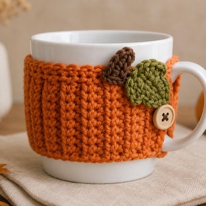 May include: A white ceramic mug featuring a textured orange crochet cozy. The cozy is decorated with a small brown crocheted stem, a green crocheted leaf, and a round wooden button with four holes, secured near the mug handle.
