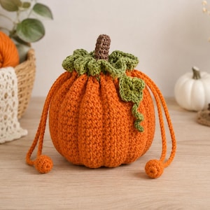 May include: A round, orange crochet pumpkin drawstring pouch featuring a brown stem, a ruffled green leaf collar, and a single green vine accent. The pouch is cinched with two orange crochet cords that end in small, round, orange crochet spheres.