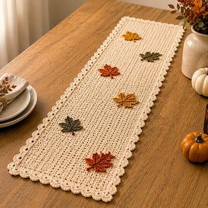 May include: A rectangular cream-colored crochet table runner featuring a scalloped edge and six small, three-dimensional fabric maple leaves in shades of orange, yellow, and dark green scattered across the center of the woven fabric surface.