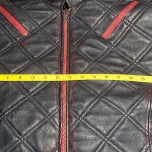 May include: A black leather jacket with a quilted diamond pattern and red zipper accents. A yellow measuring tape is stretched across the front, showing a width of approximately 27 inches from the left edge to the right edge of the garment.