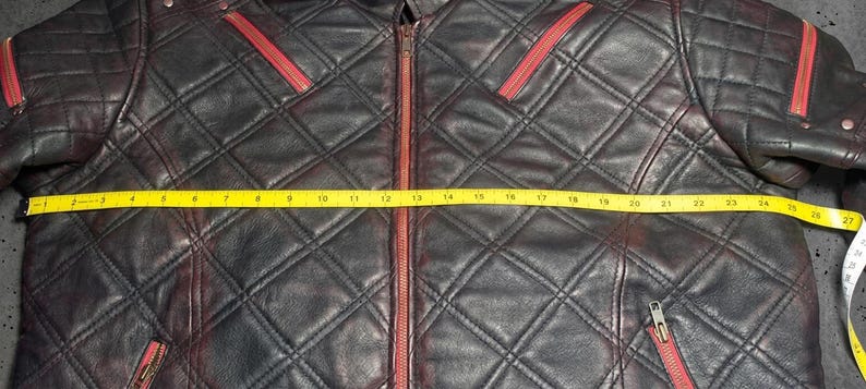 May include: A black leather jacket with a quilted diamond pattern and red zipper accents. A yellow measuring tape is stretched across the front, showing a width of approximately 27 inches from the left edge to the right edge of the garment.