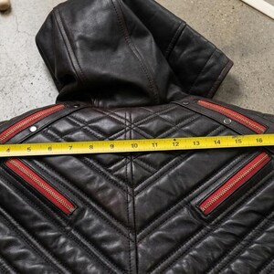 May include: A black leather hooded jacket featuring quilted paneling, red zipper accents, and silver-tone metal snaps. A yellow measuring tape is placed across the back, showing a width measurement of approximately 26 inches from edge to edge.