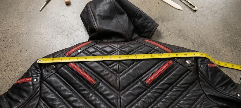 May include: A black leather hooded jacket featuring quilted paneling, red zipper accents, and silver-tone metal snaps. A yellow measuring tape is placed across the back, showing a width measurement of approximately 26 inches from edge to edge.