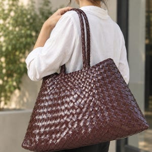 Puede incluir: Un bolso tote grande de color marrón oscuro con un intrincado patrón trenzado en toda su superficie. El bolso incluye dos correas de hombro trenzadas a juego y una base ancha y estructurada, ofreciendo un interior espacioso para llevar objetos.