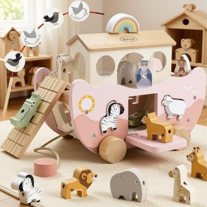 May include: A wooden Noah's Ark toy set featuring a pink and cream boat with wheels, a ramp, and a rainbow arch on top. The set includes various animal figures like a zebra, lion, elephant, giraffe, and sheep, along with two human figures.