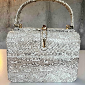 May include: A structured box-shaped handbag featuring a light grey and white snakeskin pattern. The bag includes a single top handle, a gold-tone metal clasp closure at the centre, and gold-tone hardware connecting the handle to the main body.