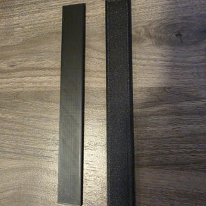 May include: Two long, narrow, rectangular strips in black. The left strip has a smooth, matte finish with visible diagonal texture lines, while the right strip features a coarser, granular, and more pronounced textured surface finish.