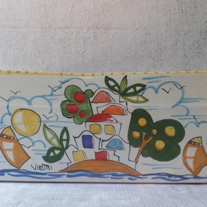 Può includere: Una pirofila rettangolare in ceramica bianca con una scena dipinta a mano di un villaggio costiero con case colorate, alberi verdi, un sole giallo e barche a vela. Il bordo presenta un motivo a pois gialli e la firma VIETRI è visibile.