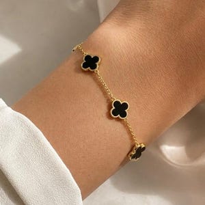 Peut inclure: Un bracelet à chaîne dorée orné de trois motifs en forme de trèfle noir avec des bordures dorées, espacés régulièrement. Les maillons métalliques délicats relient chaque motif de trèfle, formant un design continu sur le poignet.